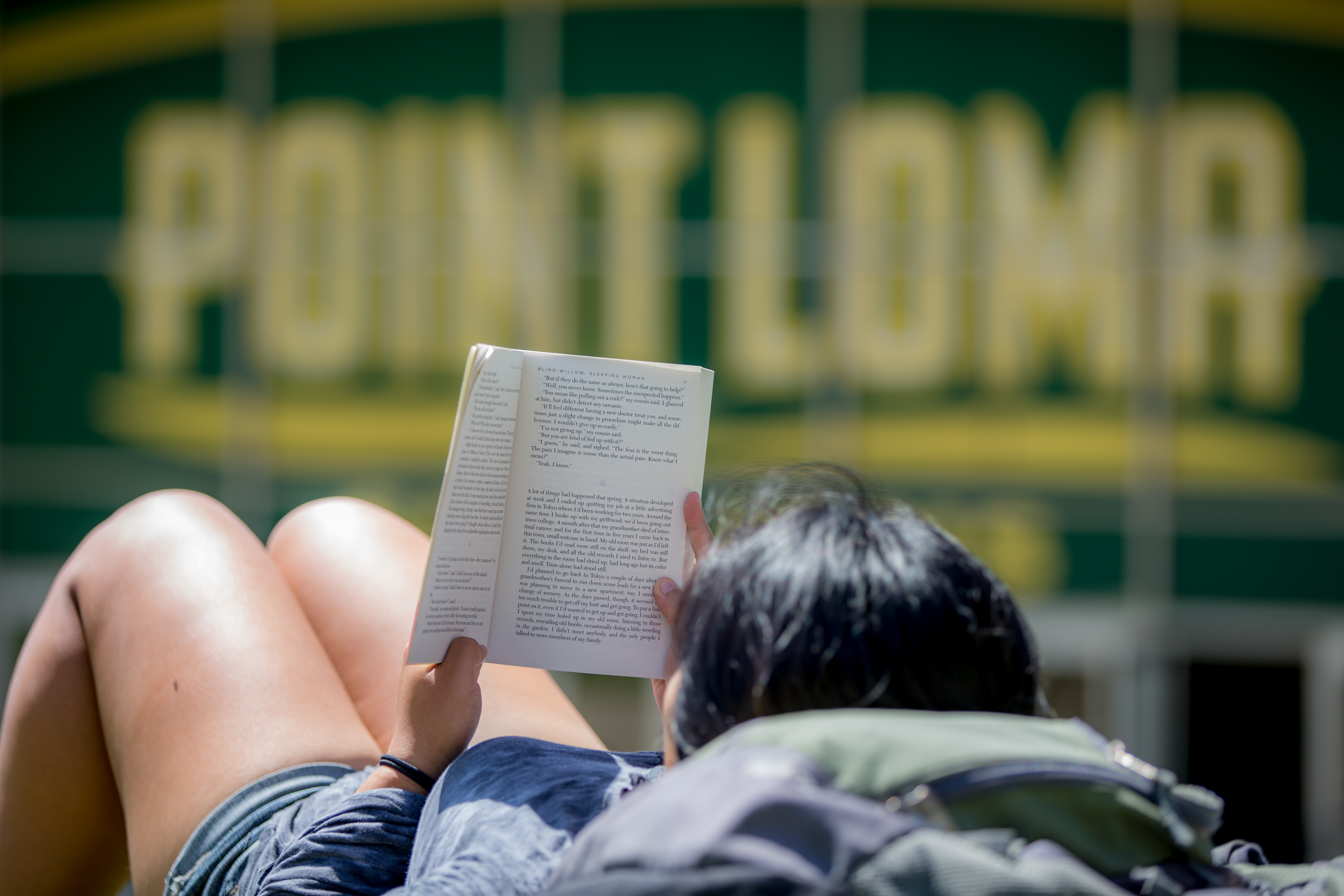 A female student with black hair is laying down outside in the sunshine. She is leaning her head on her backpack and is reading a book.