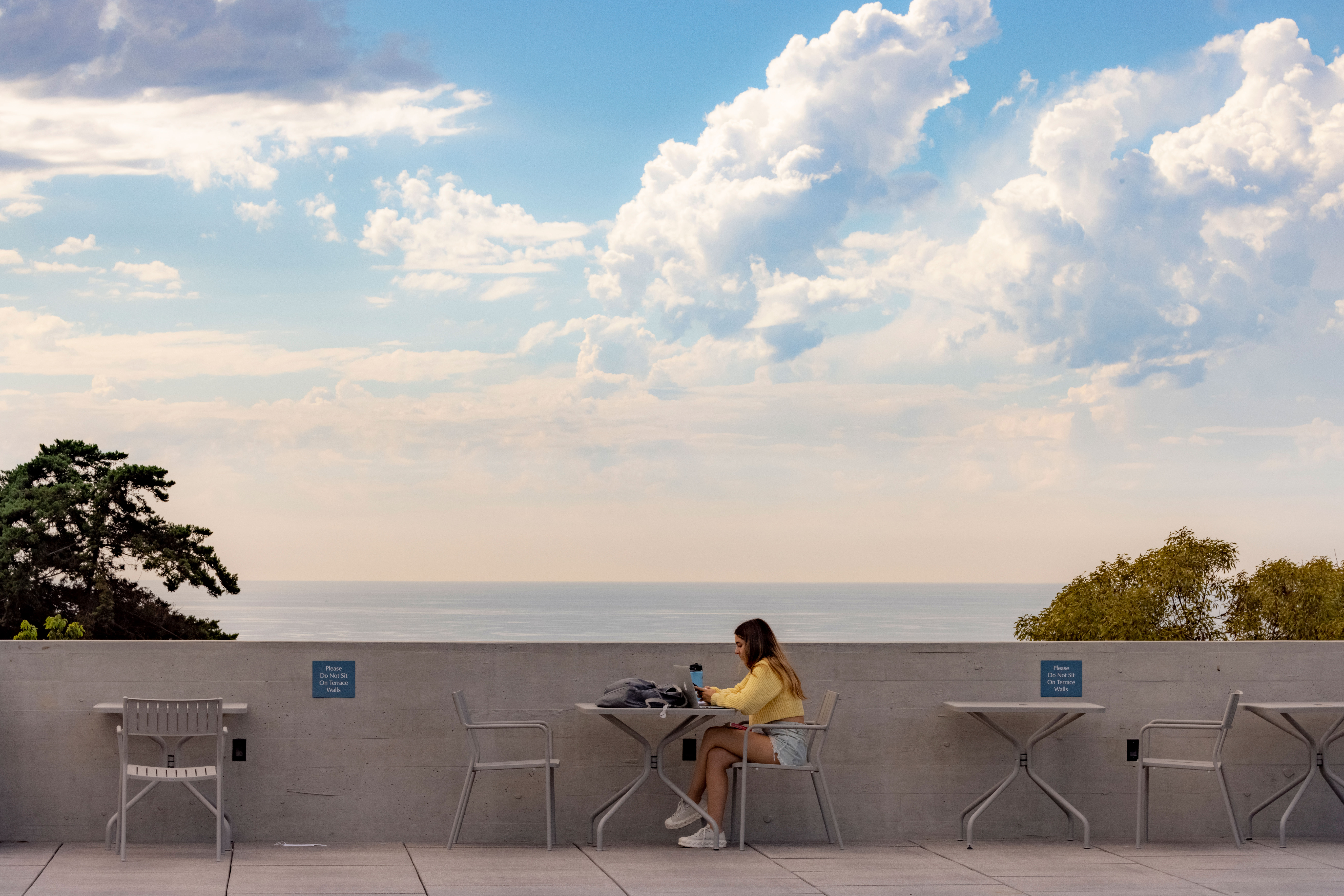 student studying at a metal table with the ocean and a bright blue sky with clouds in the background