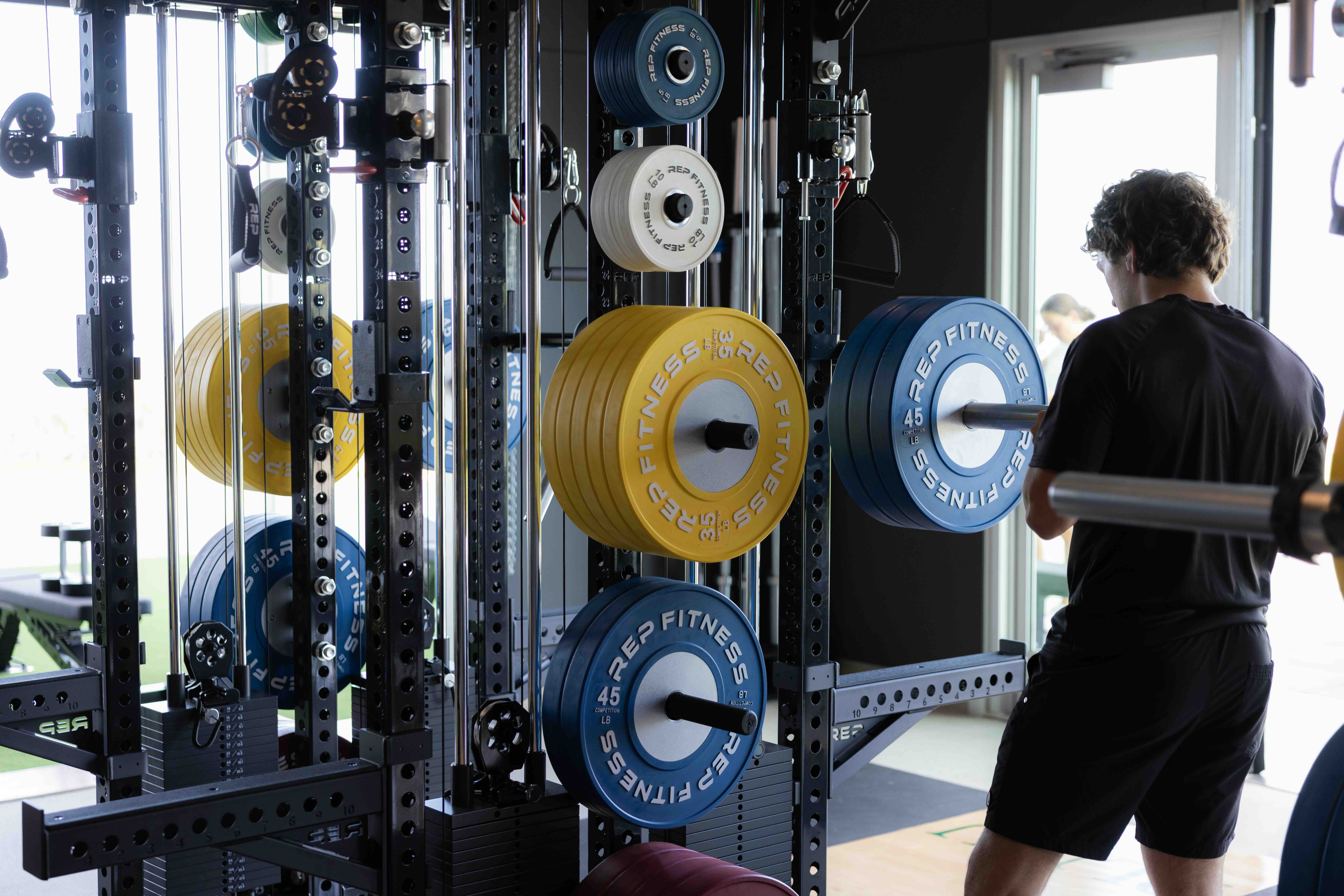 Student standing among weight racks and strength training equipment inside the Lawrence Ryan Fitness Pavilion.