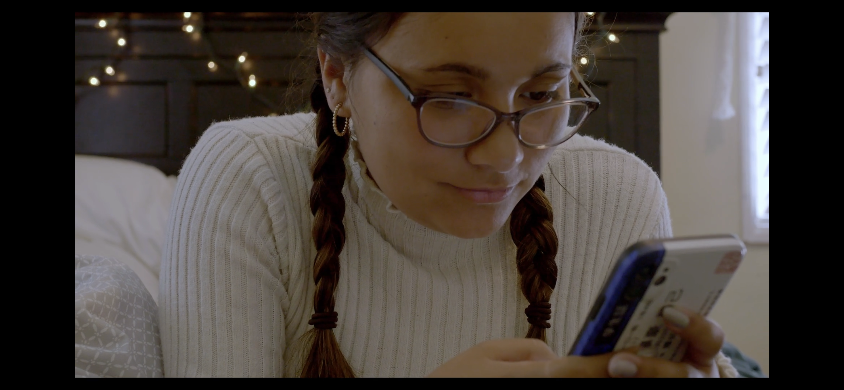 Teenage girl with braids and glasses sits on bed texting