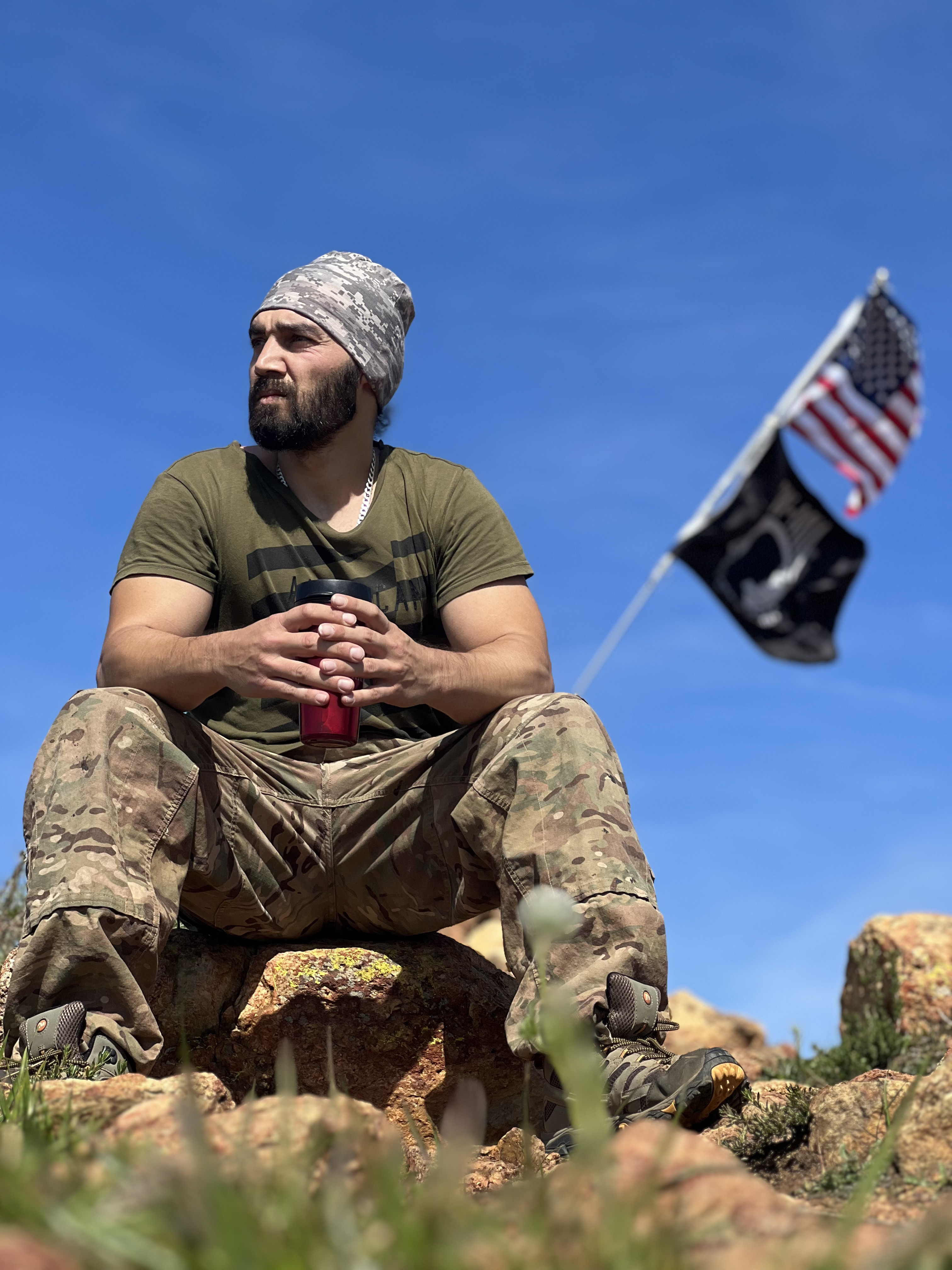 Jobe is sitting on a rock, looking to the left, and holding a red water bottle. He is wearing a green shirt and pair of pants made out of camouflage fabric.