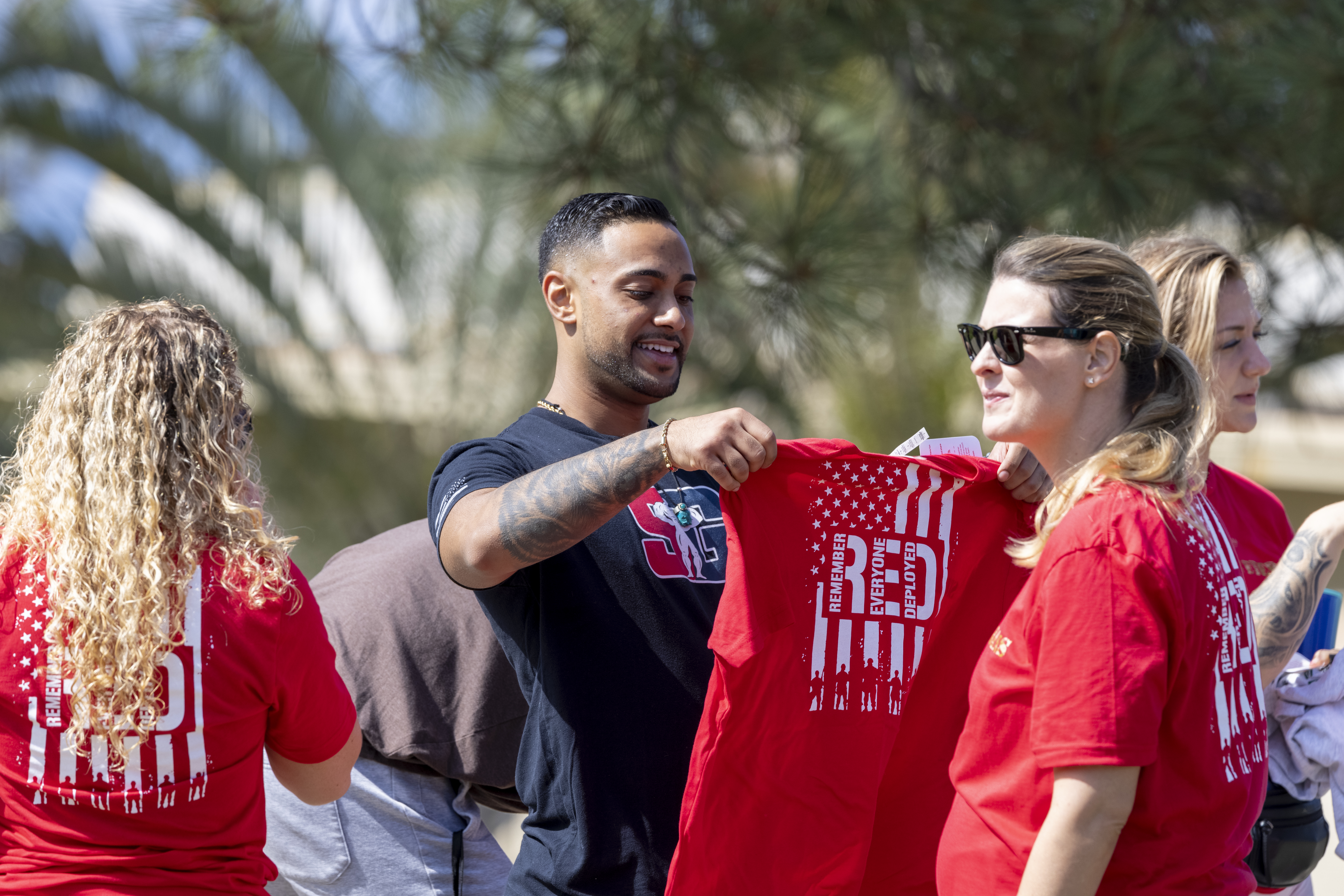 PLNU community members distributing red “Remember Everyone Deployed” shirts at an outdoor campus event.