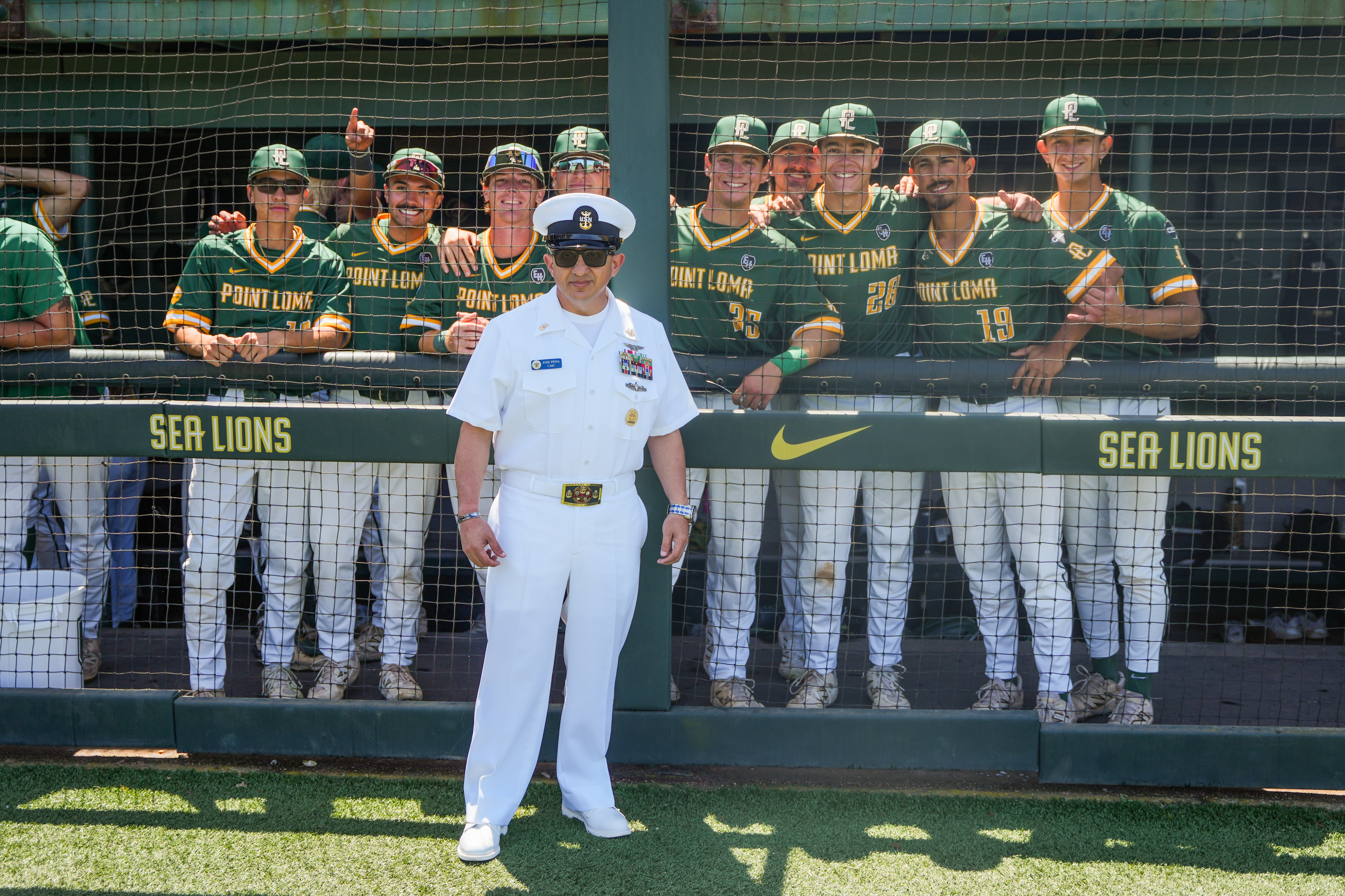 PLNU baseball players pose behind a U.S. Navy service member in uniform at the Sea Lions baseball field.