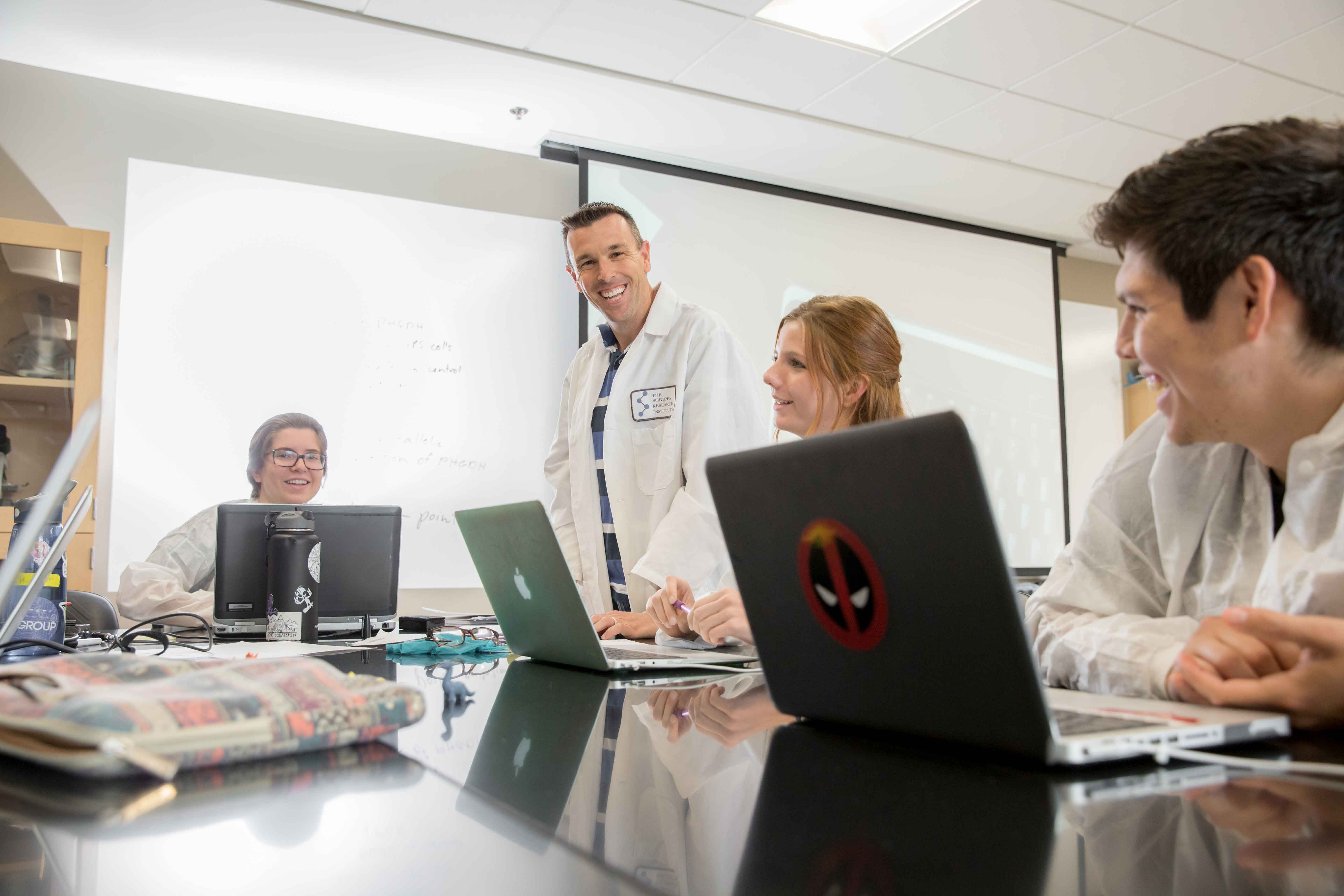 professor smiling at students looking at open laptops. everyone is wearing a white lab coat