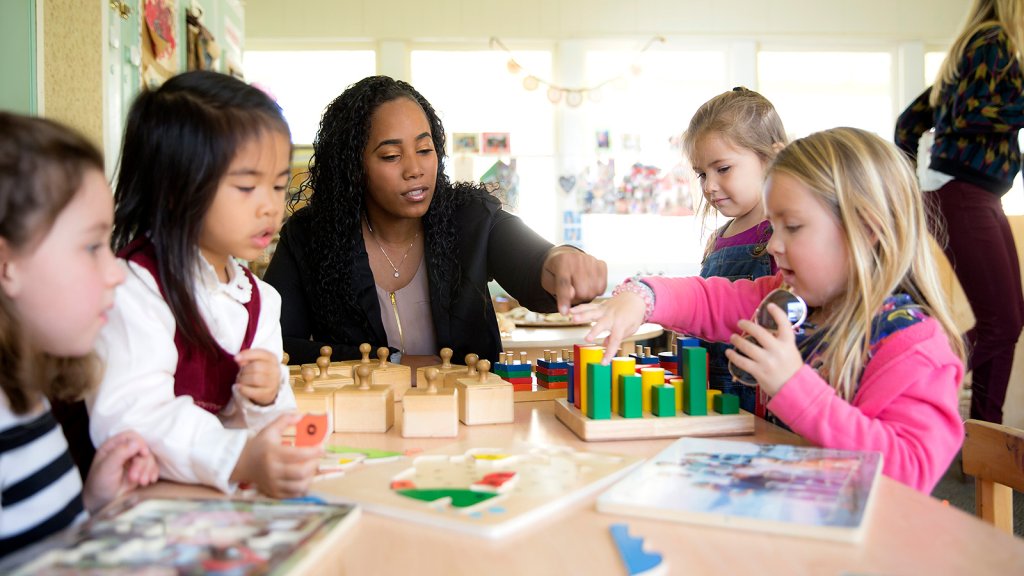 Early childhood teacher engages with small group of children in a hands-on learning activity that includes blocks and puzzles in a classroom.