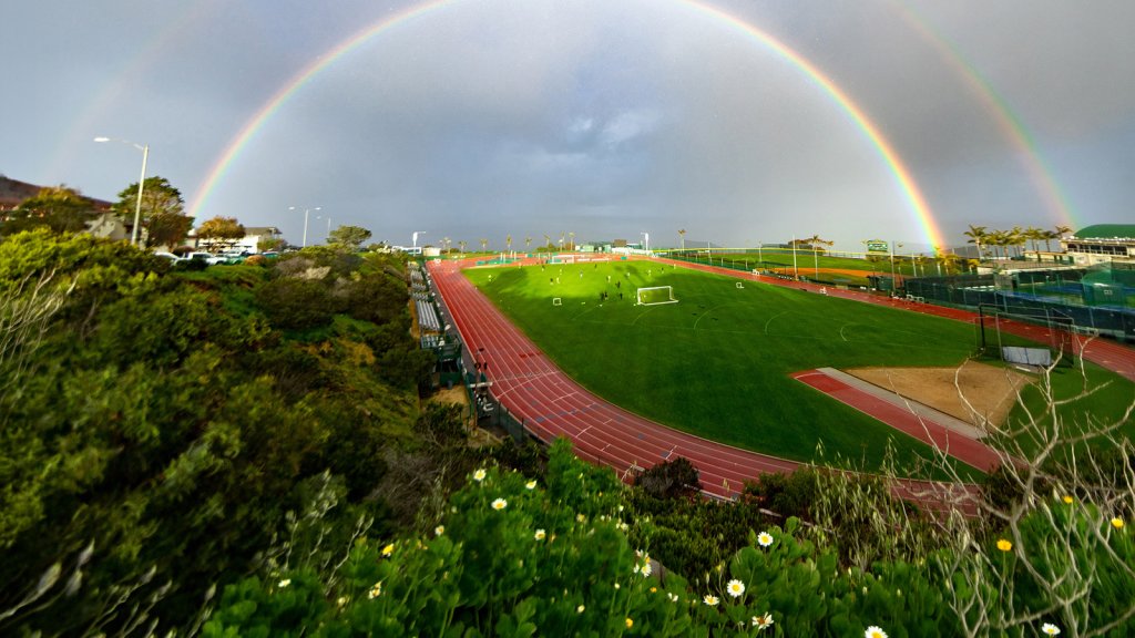 Image of university's track and field where a triple rainbow is seen in cloudy sky following rainy weather.