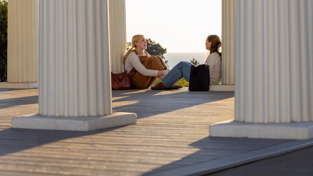 Two PLNU students sit resting along columns while conversing at the Greek Amphitheatre with the Pacific Ocean in the background.