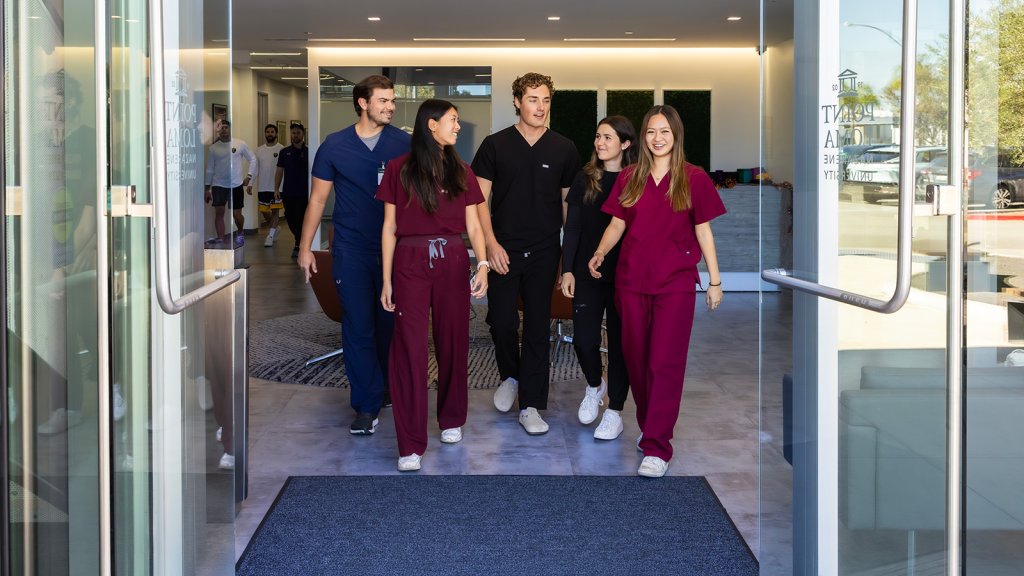 PLNU nursing students dressed in scrubs connecting in conversation while walking through entrance doors of Balboa Campus.