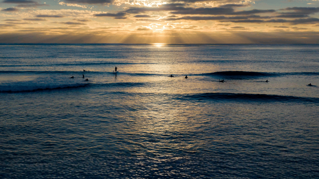 Surfers wait for waves as sunlight breaks through clouds over the Pacific Ocean.