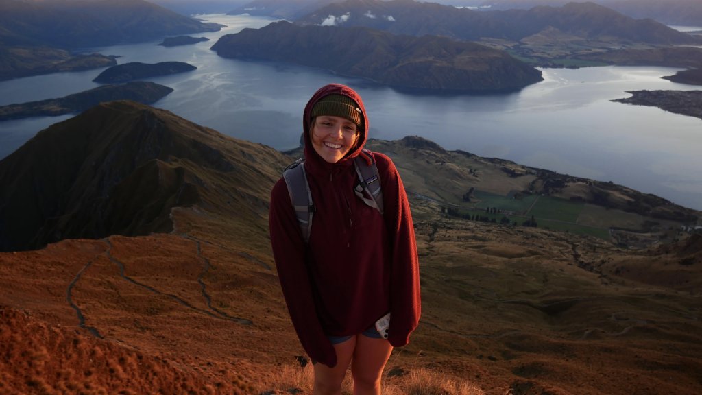 PLNU student wearing a backpack stands on a mountain overlooking a winding lake and mountain landscape at sunrise in New Zealand.