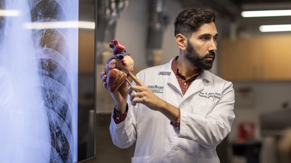 Dr. Joy Y. Balta, wearing a white lab coat, points to a model of a human heart in front of a large medical screen displaying an X-ray image. He appears to be in an educational setting, explaining anatomy concepts to an audience.