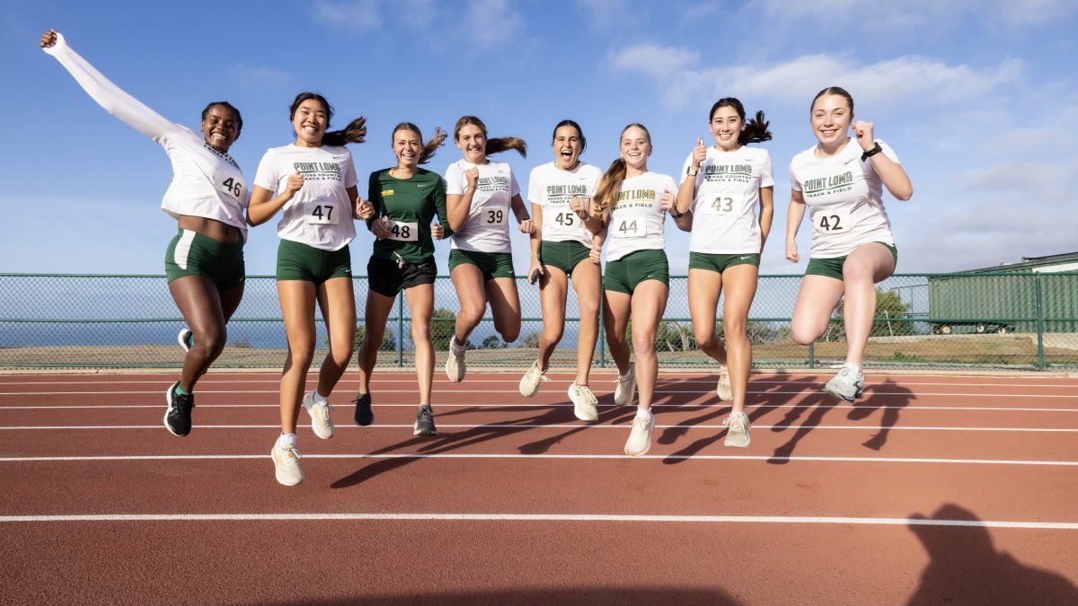Female PLNU athletes all jump in celebration at a track meet on the university track with the Pacific Ocean in the background.