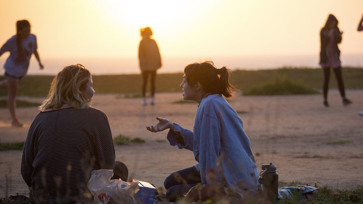 Two girls sit on the grass at sunset discussing life while others play baseball in the background.