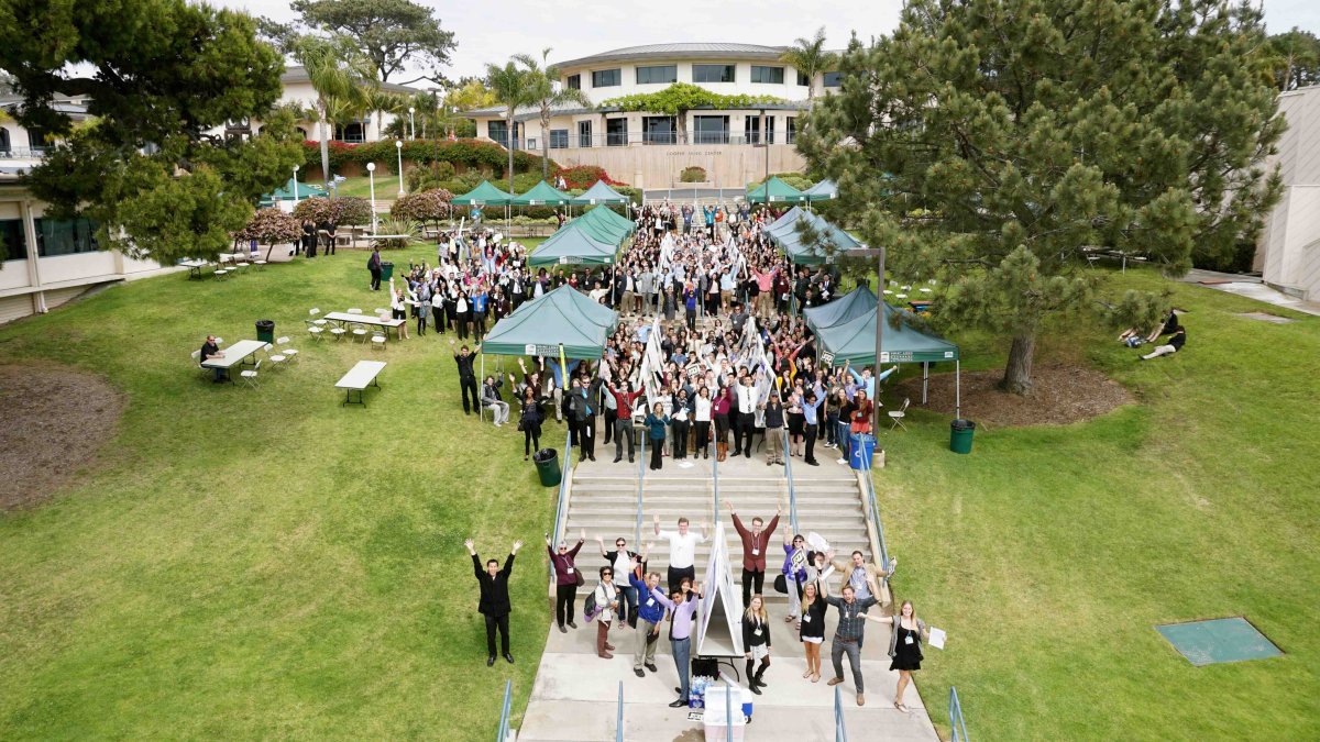 Large group of students, faculty, and research presenters gather outdoors on the PLNU campus during an academic research event.