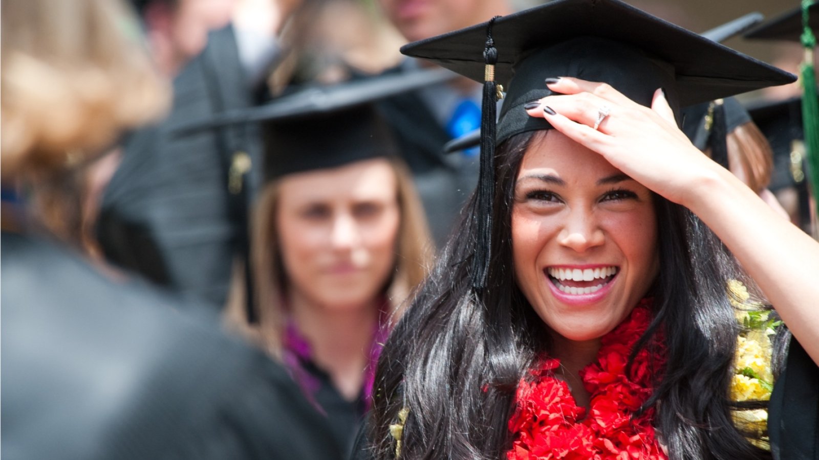A female student in graduation cap, gown, and red flower lei excitedly congratulates other students