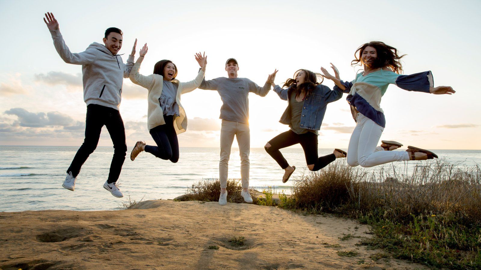 Small group of students jump in air with sunset and ocean behind them