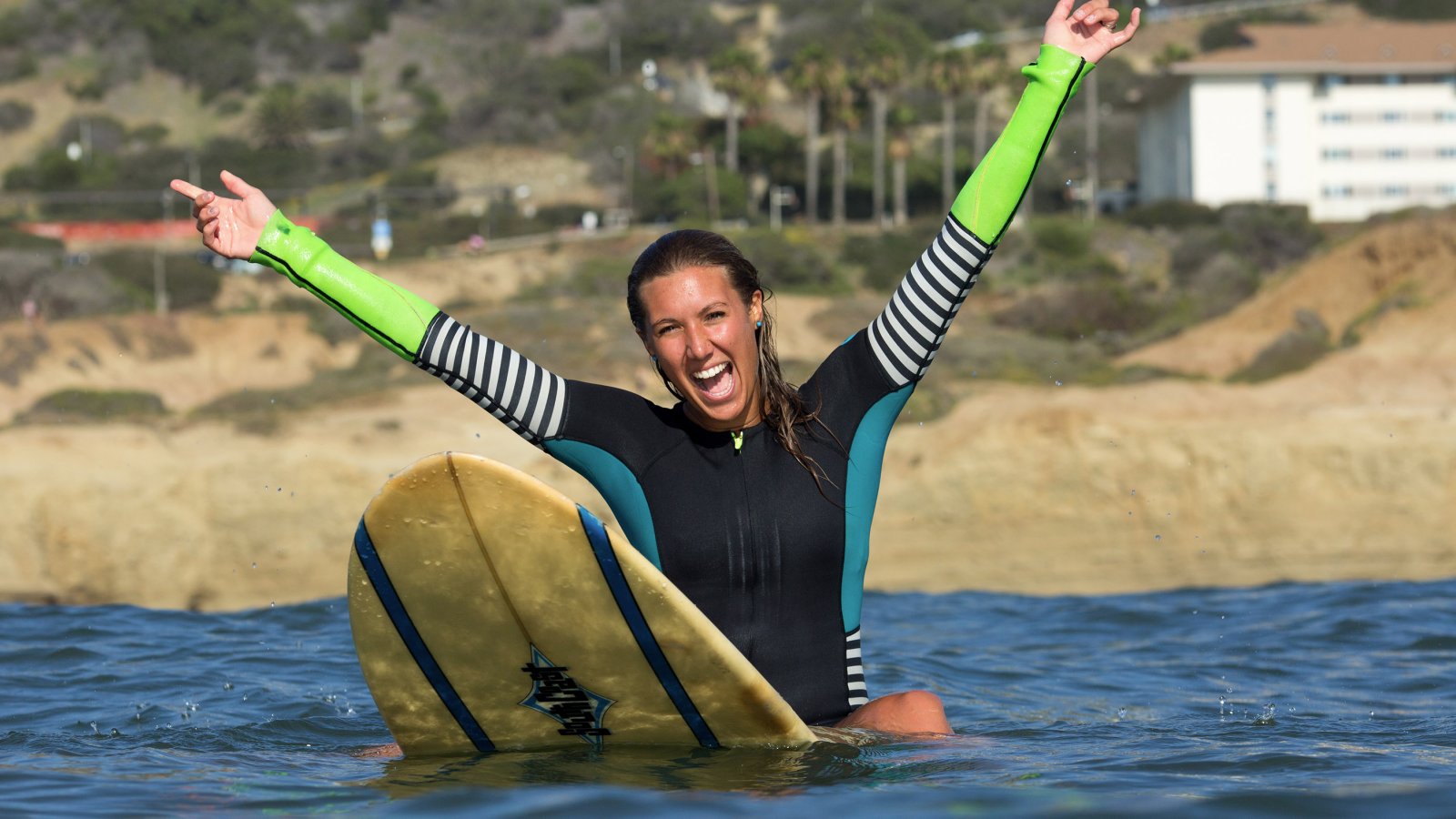 Young woman wearing a wetsuit sits on a surfboard in the ocean with her hands above her head in excitement