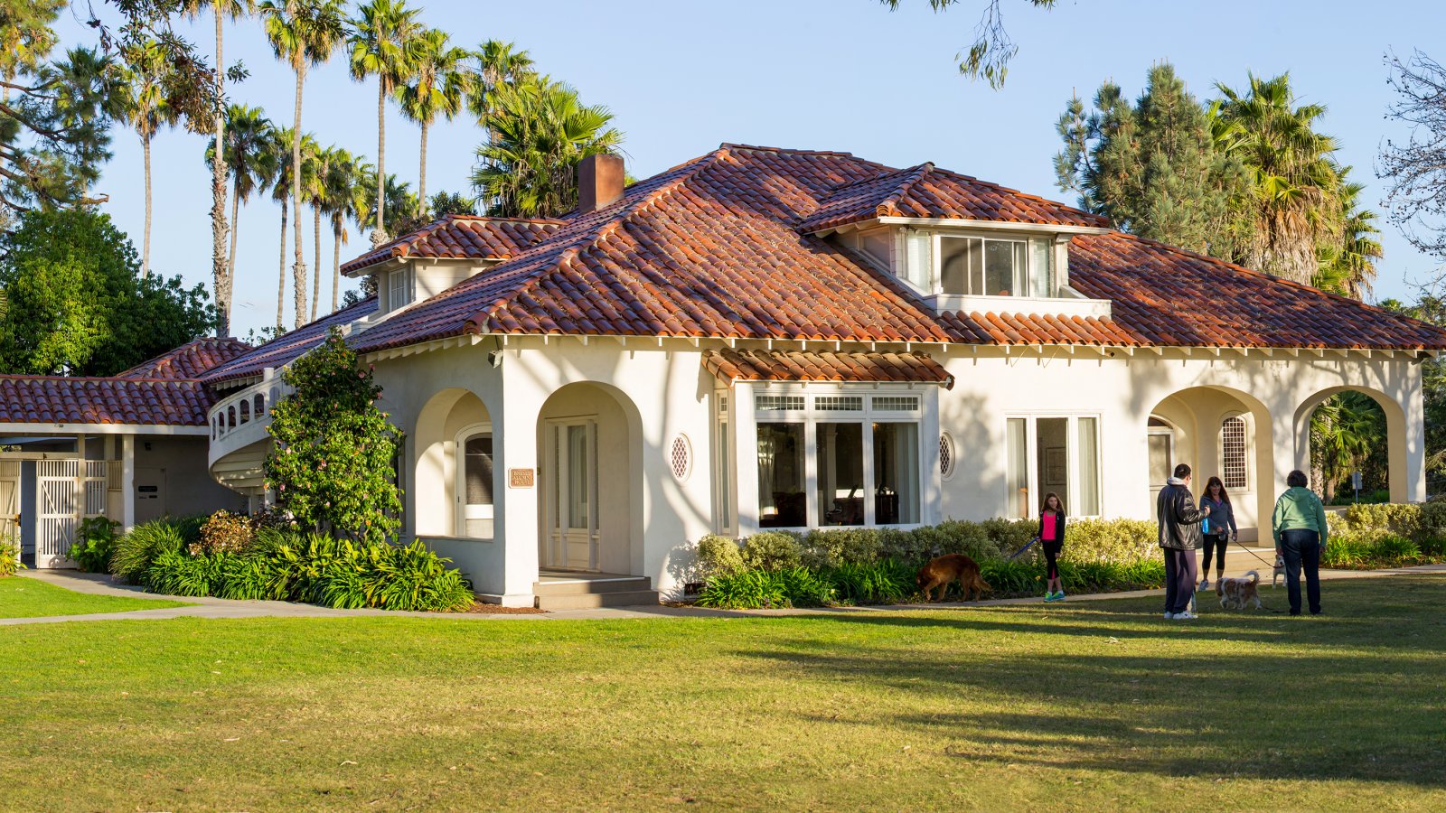A family walks their dogs on alumni lawn with PLNU's Alumni Office in the background.