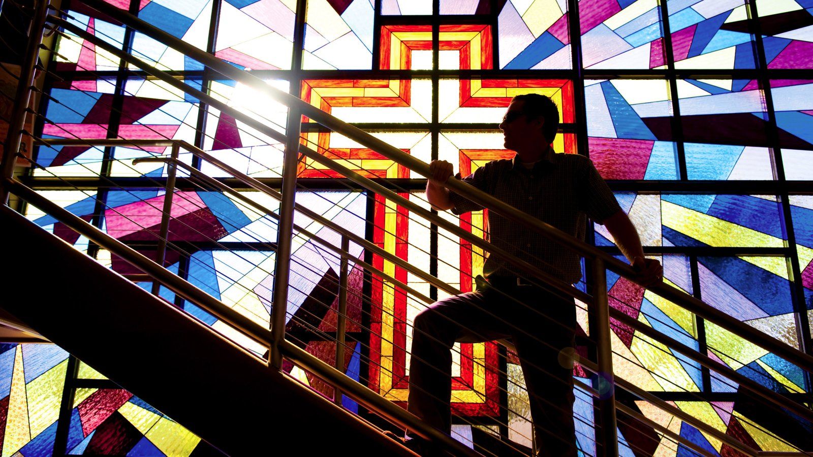Man climbs staircase in shadow, backlit by sun coming through giant stained glass window of a cross
