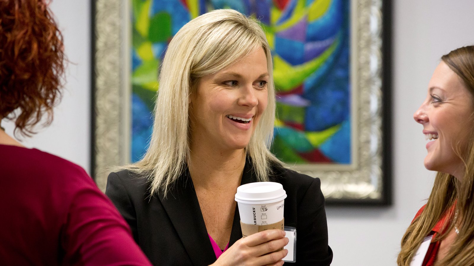 Professor Lindsey Lupo holds a coffee and talks to a female student while standing in front of a painting