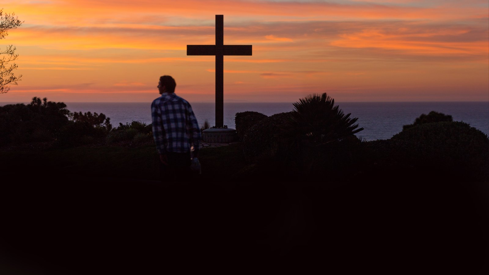 Male student walks past the cross with view of a sunset.