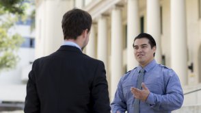 A man who works in public administration is standing outside of San Diego City Hall. He is wearing a blue button down and is talking to a man wearing a black suit.