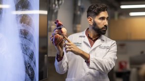 Dr. Joy Y. Balta, wearing a white lab coat, points to a model of a human heart in front of a large medical screen displaying an X-ray image. He appears to be in an educational setting, explaining anatomy concepts to an audience.
