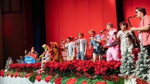 several college students singing into microphones, wearing christmas outfits and beach outfits. The backdrop is red and the stage is lined with red poinsettia flowers.