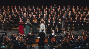 large choir with 7 featured singers in front. The conductor and orchestra are below the choir. 