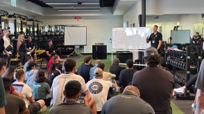 A group of students and professionals sit on turf flooring inside a modern training facility, listening to a speaker standing beside a whiteboard filled with notes. Weight racks, barbells, and gym equipment line the room as the presenter gestures while leading an instructional session.