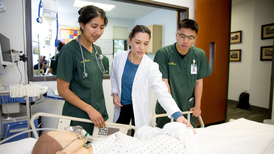 Two student nurses in green scrubs stand on each side of a nurse in a white coat. The three nurses are looking at a manikin laying on a hospital bed.