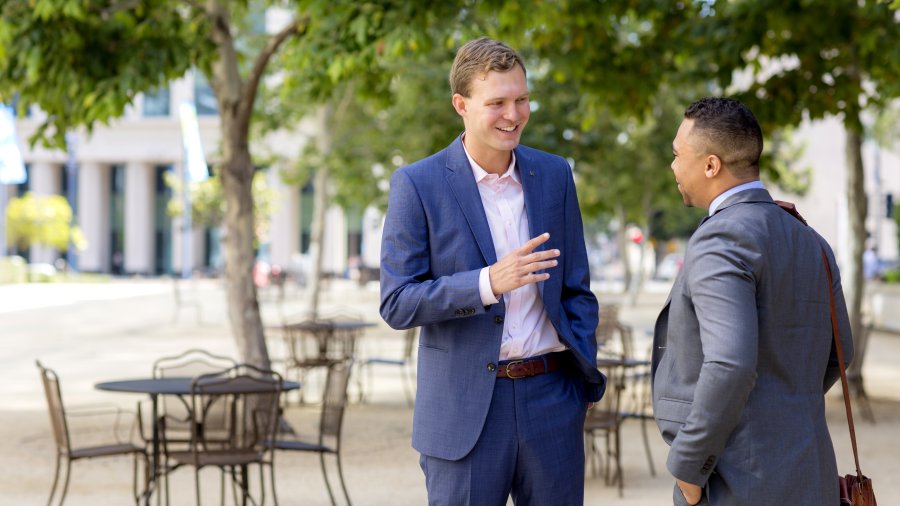 Two business men wearing suits are standing outside on a patio. They are talking and smiling.