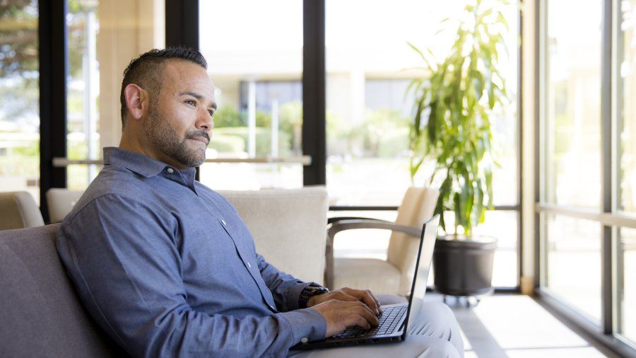 A man in a blue button down and grey pants sits on a couch with a laptop on his lap. He is looking up, thinking whether he should pursue a Master's in Writing or an MFA in Writing.