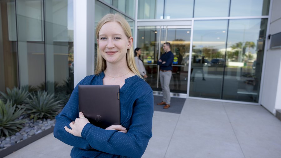 A female grad student is wearing a blue sweater and is standing outside of her graduate program's building. She is smiling and holding her laptop to her chest.