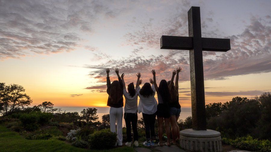 Students raise their hands next to the PLNU cross