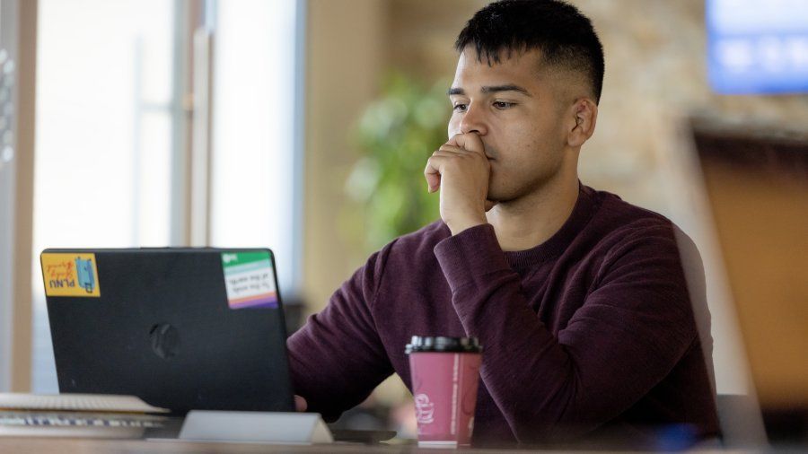 A male student is sitting at a desk and working on a graphic design project on his laptop.
