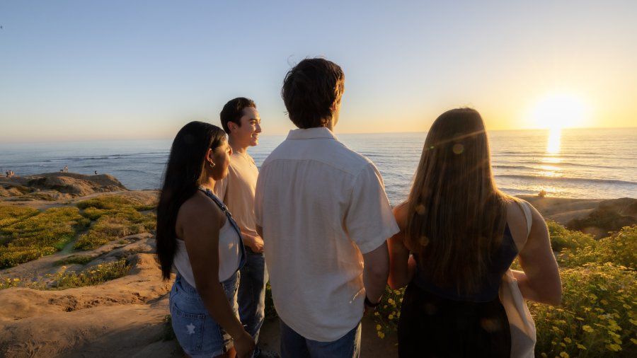 Four students stand on Sunset Cliffs together. They are smiling and looking at the sunset in the horizon.