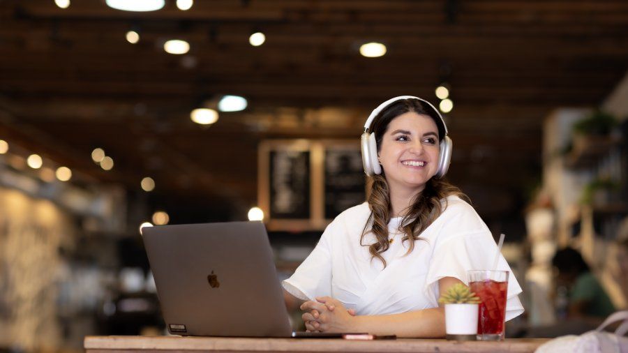 A woman is smiling and sitting in a coffee shop. She is wearing headphones and has an open laptop in front of her.