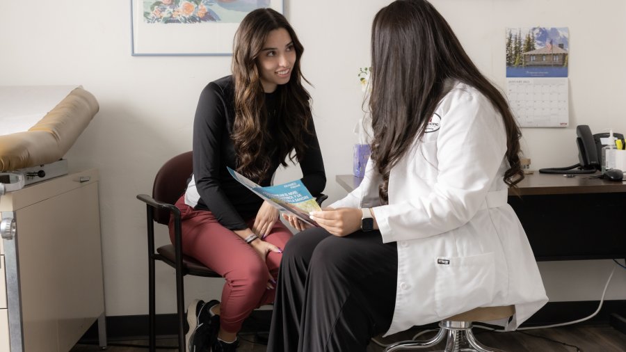 Two nurses sit in an exam room. One is holding a medical pamphlet while they are talking to each other.