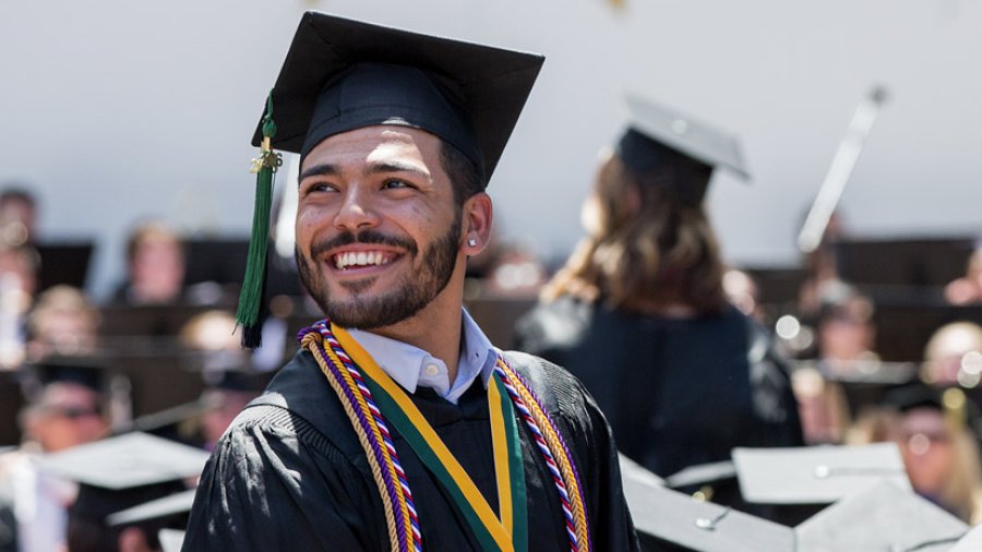 A young man stands smiling and looking into the distance wearing a cap and gown