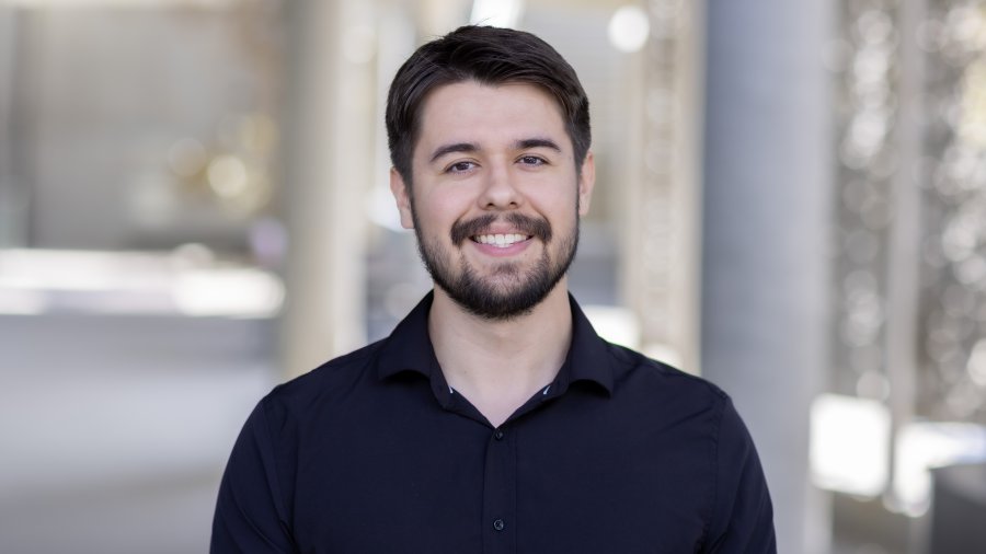 A smiling man with short brown hair and a beard wearing a black collared shirt poses for a professional portrait.
