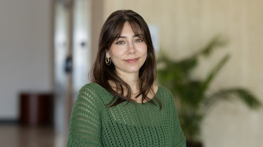 A woman with shoulder-length brown hair wearing a green knit top and gold hoop earrings poses for a professional portrait.