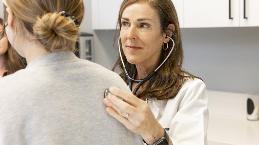 A female nurse with brown hair is holding a stethoscope to her patient's back.