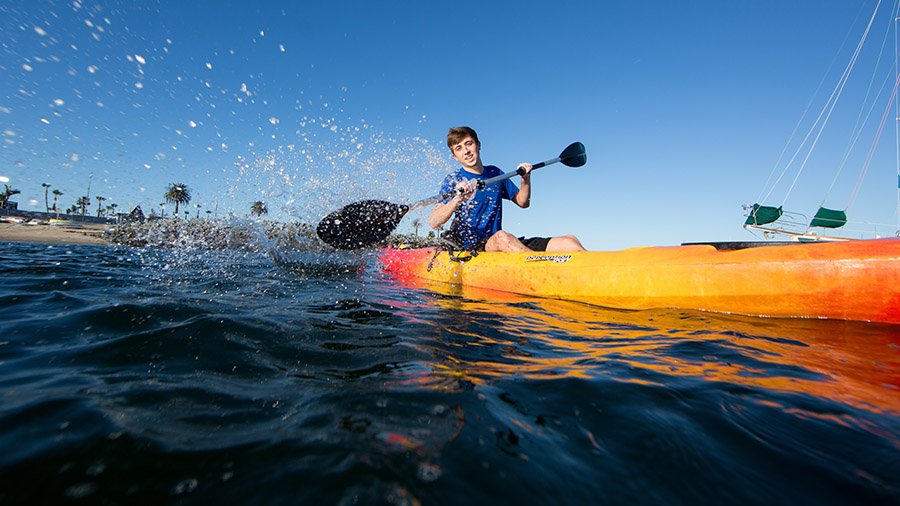 A student splashes water at the camera while paddling a kayak off Shelter Island.