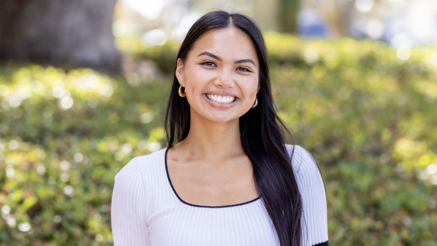 A smiling woman with long dark hair wearing a white top with black trim and gold hoop earrings poses outdoors in front of greenery.