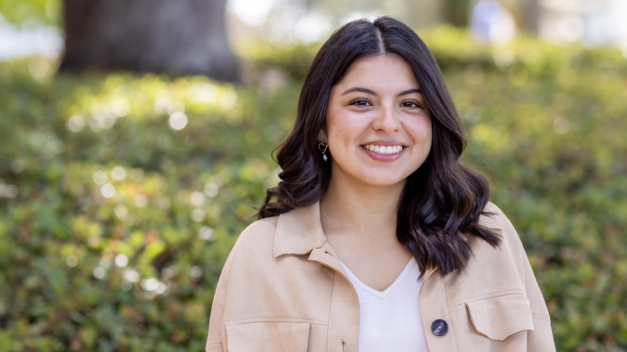 A smiling woman with shoulder-length dark hair wearing a tan jacket and white top poses outdoors in front of greenery.