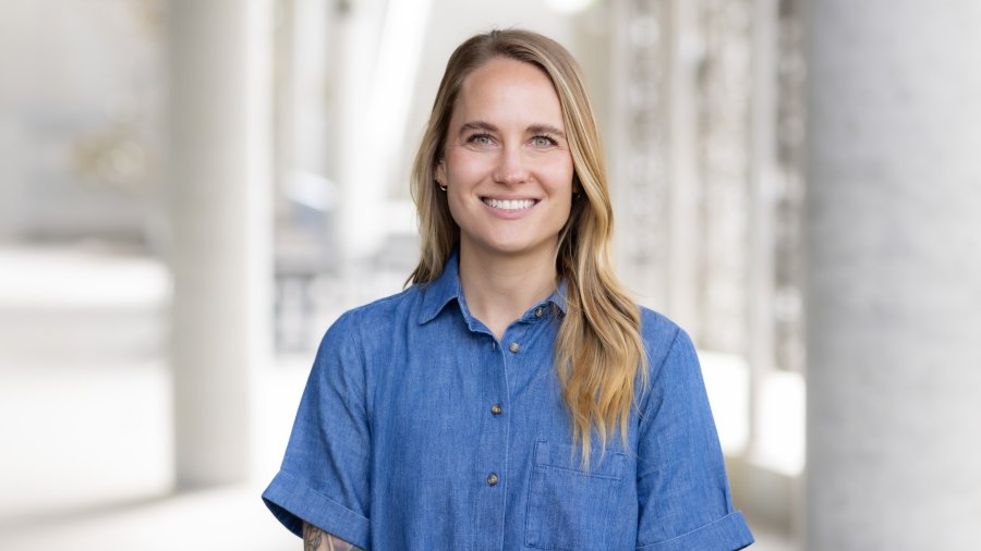 A smiling woman with long blonde hair wearing a blue button-up shirt poses for a professional portrait.