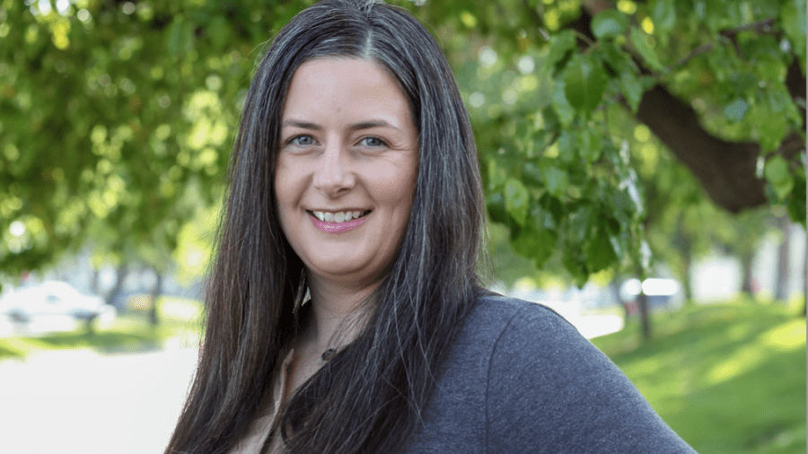 A smiling woman with long dark hair wearing a gray cardigan poses outdoors beneath a leafy green tree.