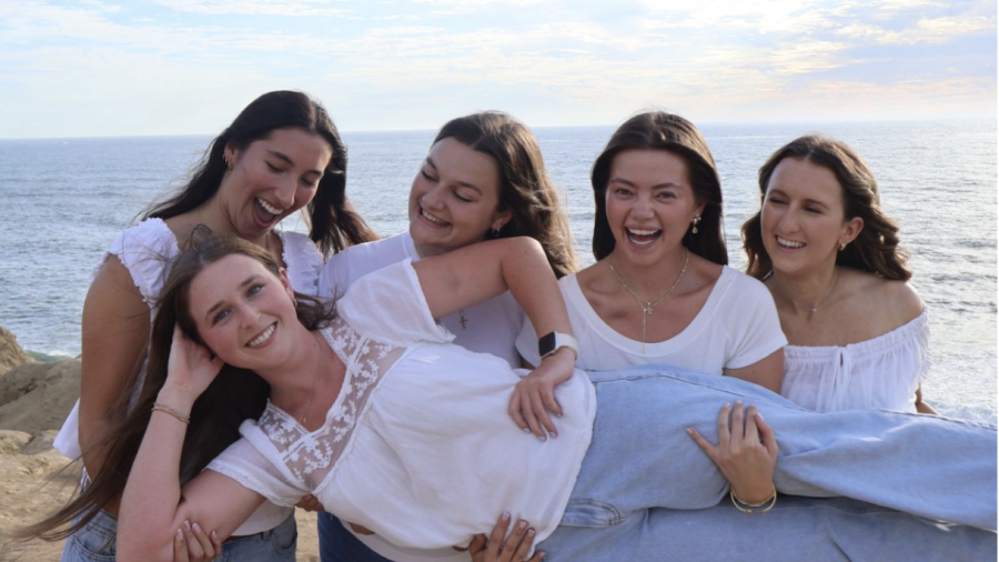 Five smiling women dressed in white tops and blue jeans pose playfully on a coastal cliff overlooking the ocean, with four holding one woman horizontally beneath a partly cloudy blue sky.