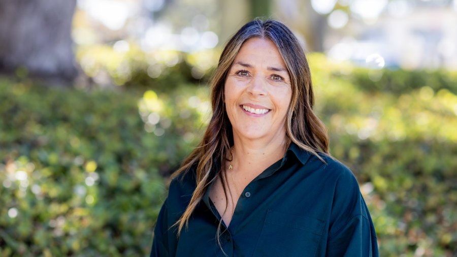 A smiling woman with long dark hair wearing a dark teal button-up shirt poses outdoors in front of greenery.